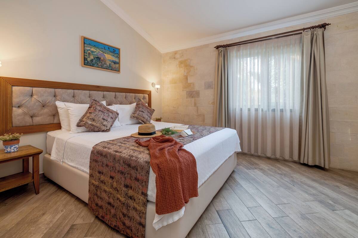 Quarto com uma cama grande, mesa de cabeceira, luminária na parede, janela com cortinas e um quadro na parede.