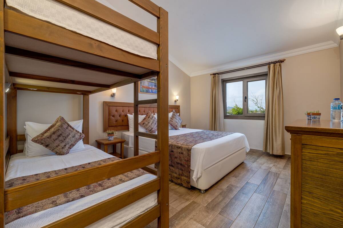 Quarto de hotel com duas camas, cabeceiras de madeira, uma janela grande com cortinas, mesa de cabeceira com abajur e um dispensador de água.