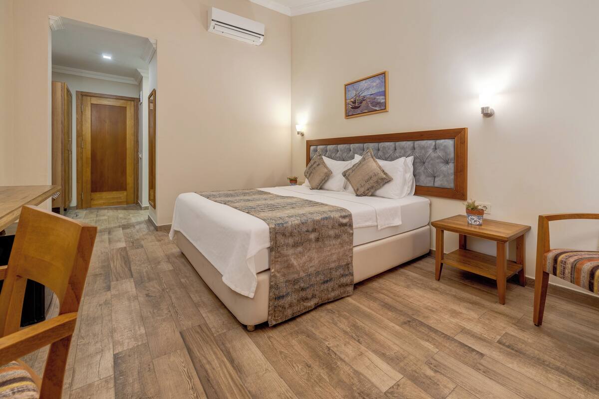 Quarto de hotel com cama, uma cadeira de madeira, uma mesinha e um quadro na parede.