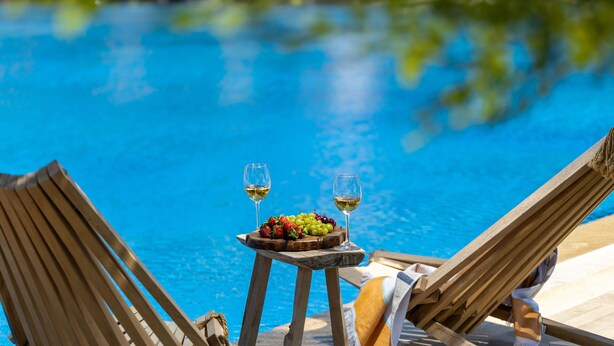 Una silla de lounge de madera junto a una piscina, con una mesa que sostiene un plato de fruta y dos copas de vino blanco.