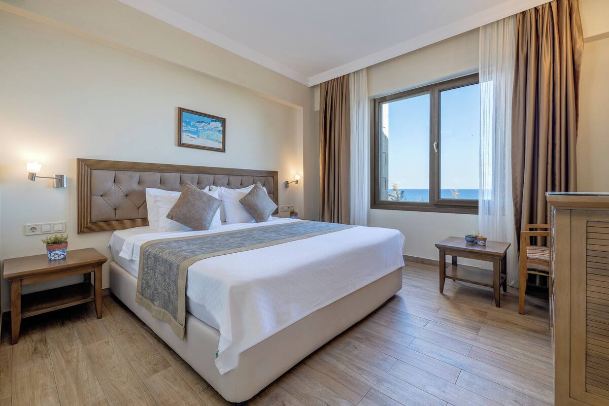 A hotel room with a large bed, bedside tables, a view of the sea, and a painting on the wall.