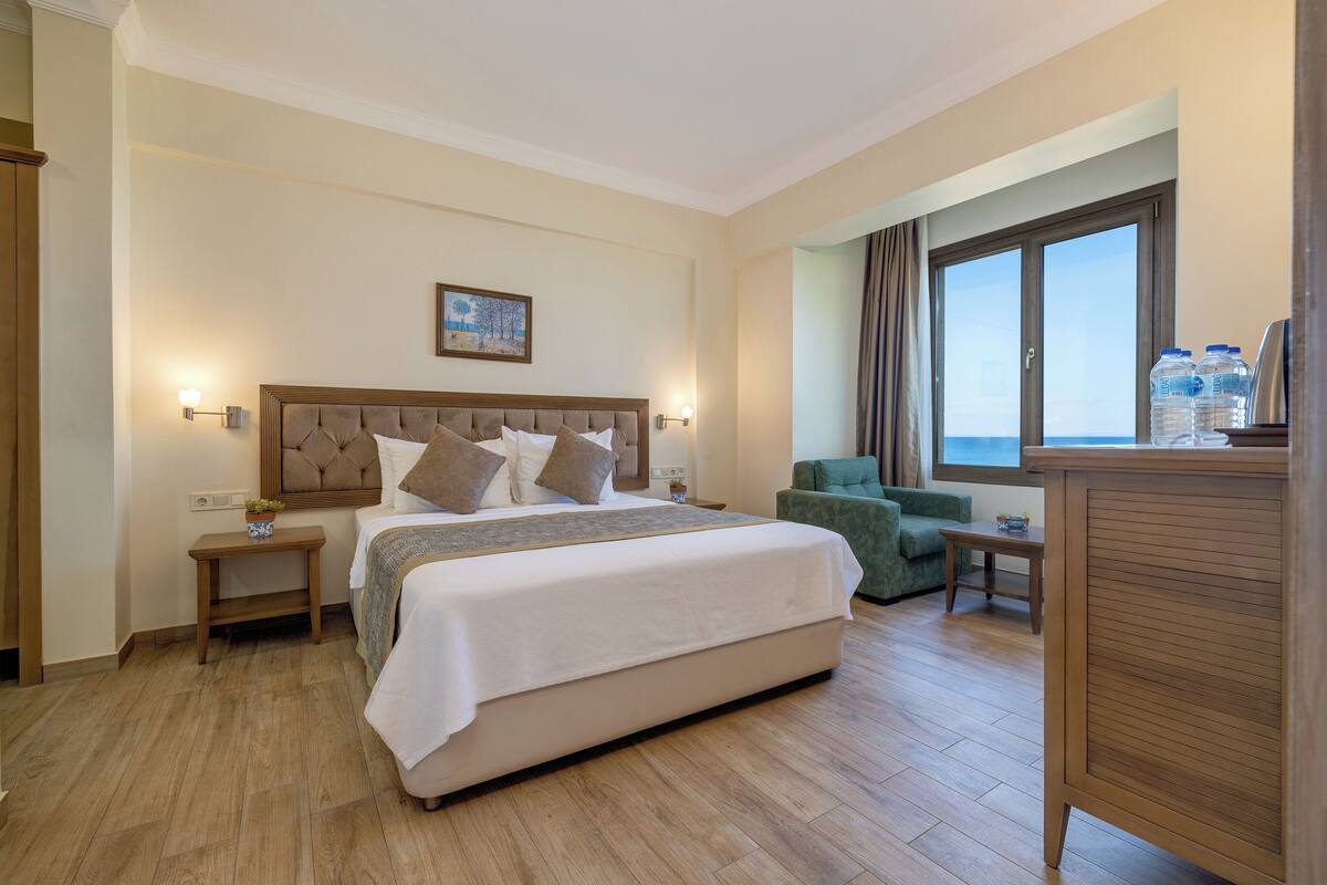 A hotel room with a large bed, a green armchair, a small table, and a view of the sea through a window.