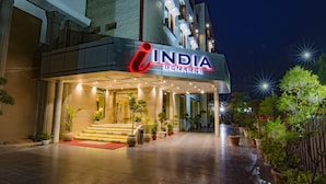 A hotel entrance with a sign reading "INDIA BENARES," featuring a modern design with a red and white logo, potted plants, and a red carpet leading to the entrance. - The India Benares (Varanasi)