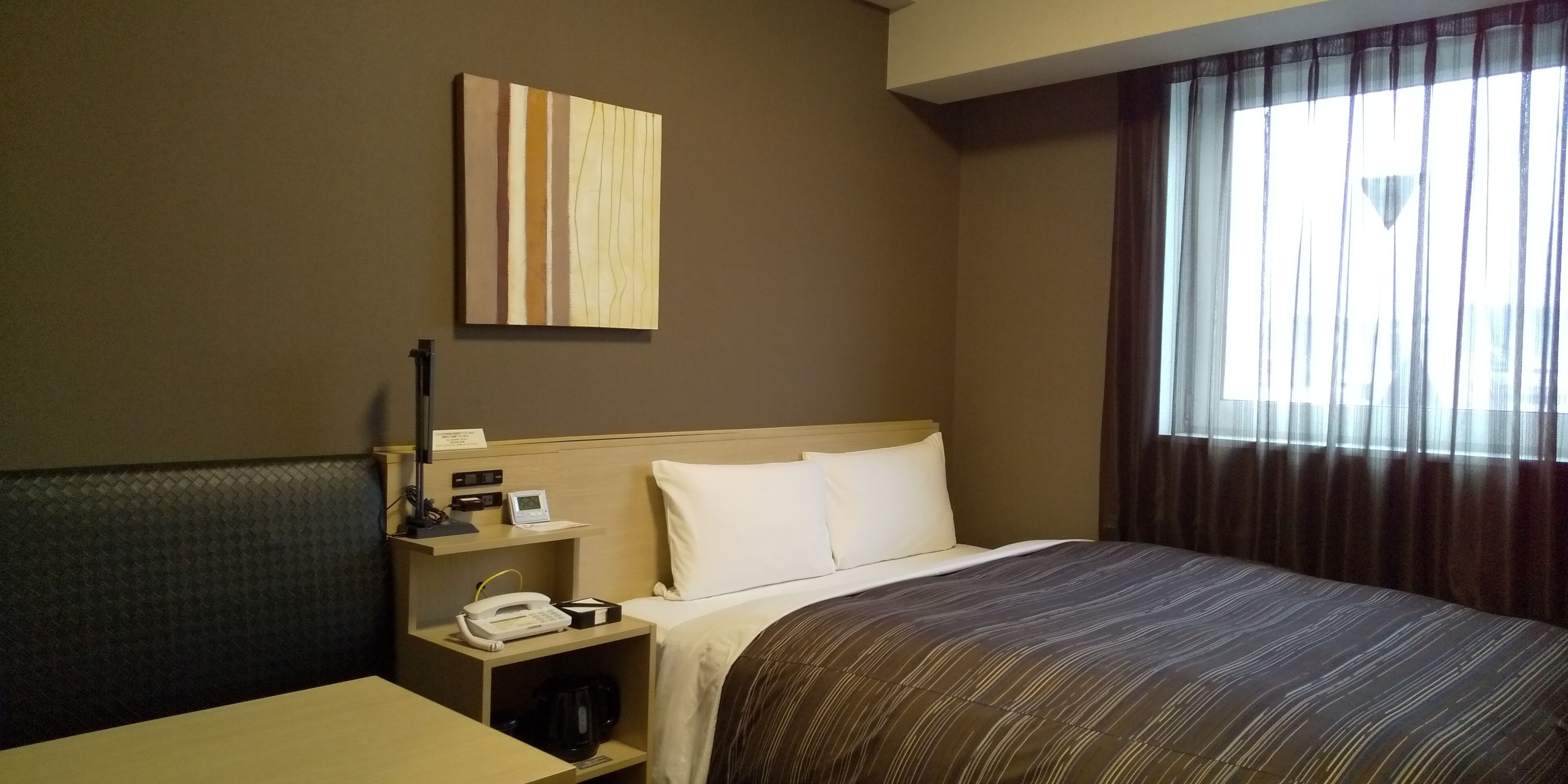 A hotel room with a bed, bedside table, lamp, phone, and a painting on the wall.