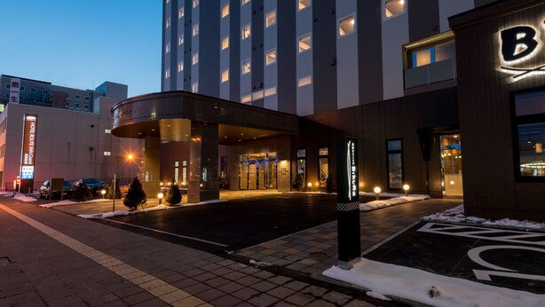 A hotel entrance with a modern design, featuring a large building with multiple windows and a prominent 'B' sign. - Hotel Route-Inn Grand Kitami Ekimae (Kitami)