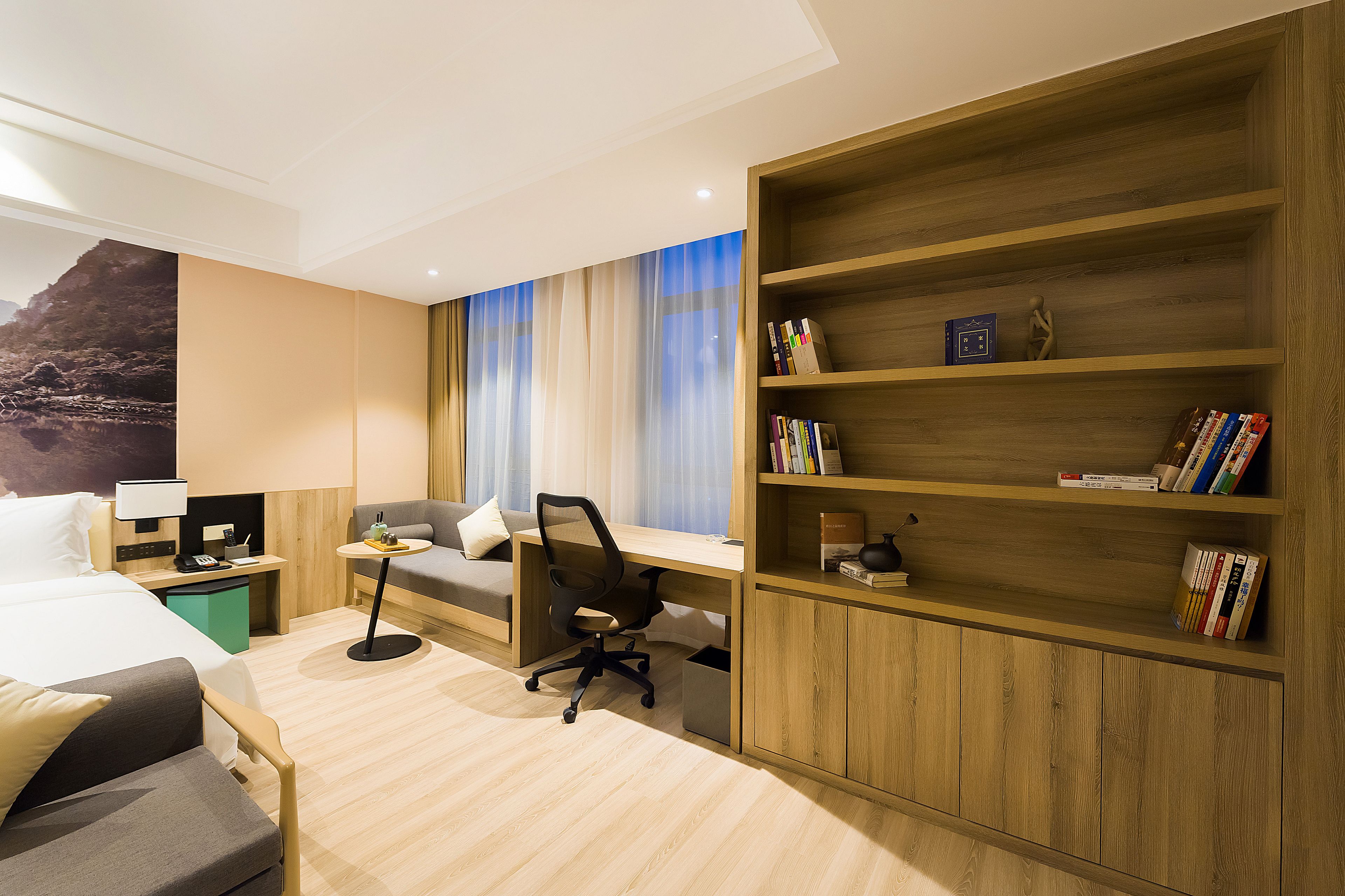 Ein modernes Hotelzimmer mit Bett, Schreibtisch, Stuhl, Bücherregal und einem großen Fenster mit Vorhängen.