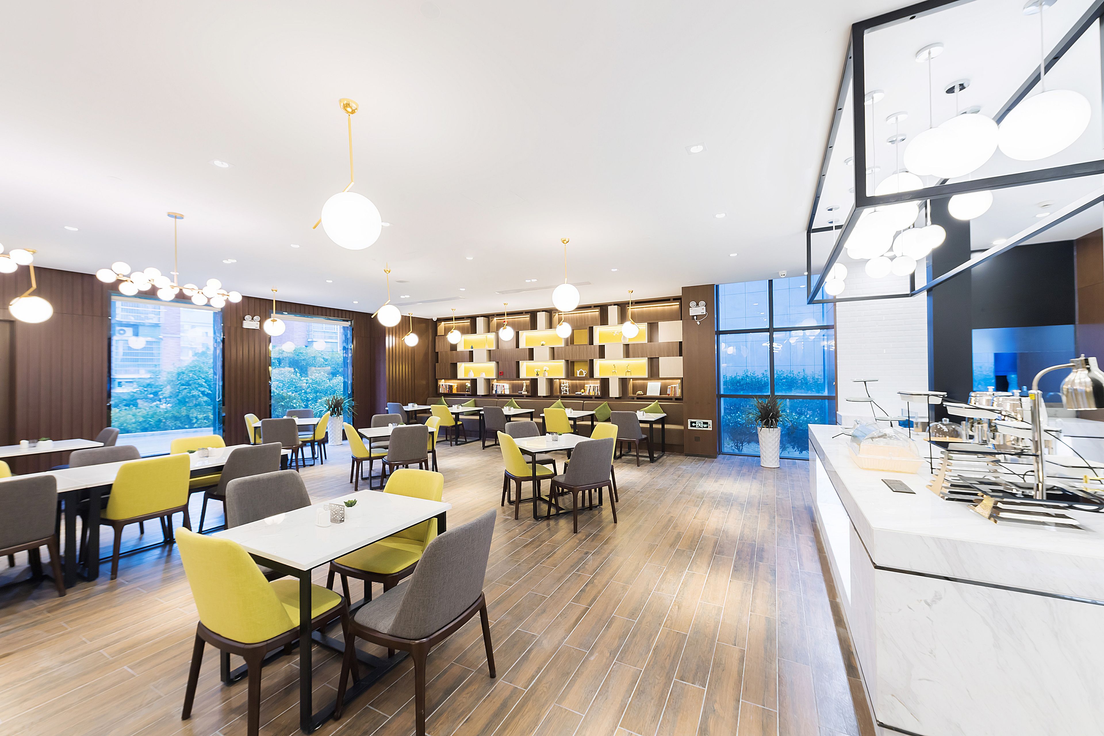 Un comedor moderno con varias mesas y sillas, una encimera de cocina y ventanas amplias que ofrecen vistas al exterior.