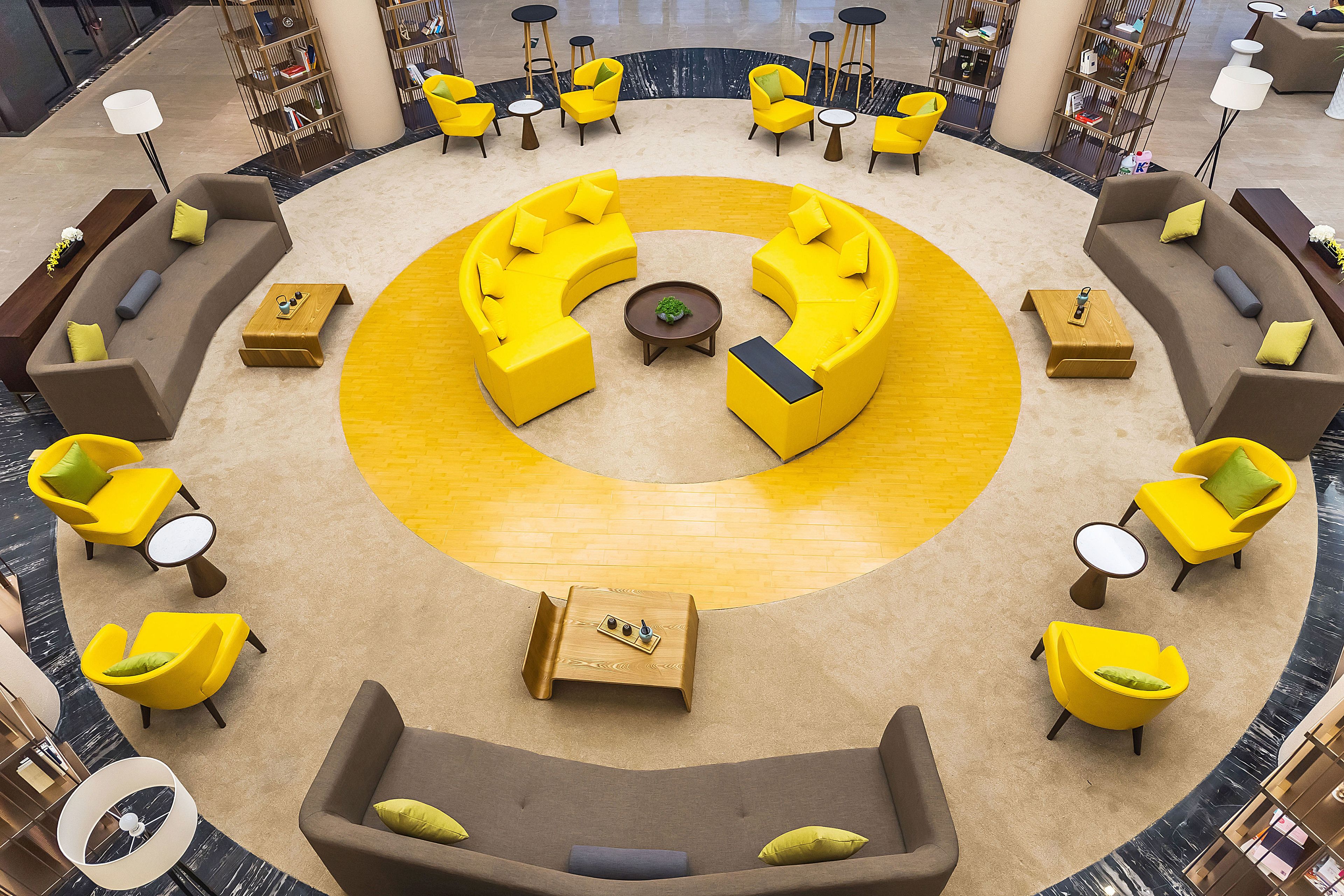 Un área de estar circular con sofás amarillos y marrones, rodeada de mesitas y sillas, en un vestíbulo moderno con una mesa redonda central.