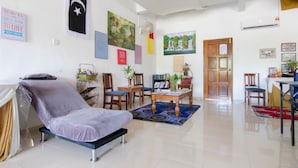 Un salon comprenant un canapé, des fauteuils, une table basse et un coin repas avec une table et des chaises. Les murs sont décorés et une porte donne accès à une autre pièce.