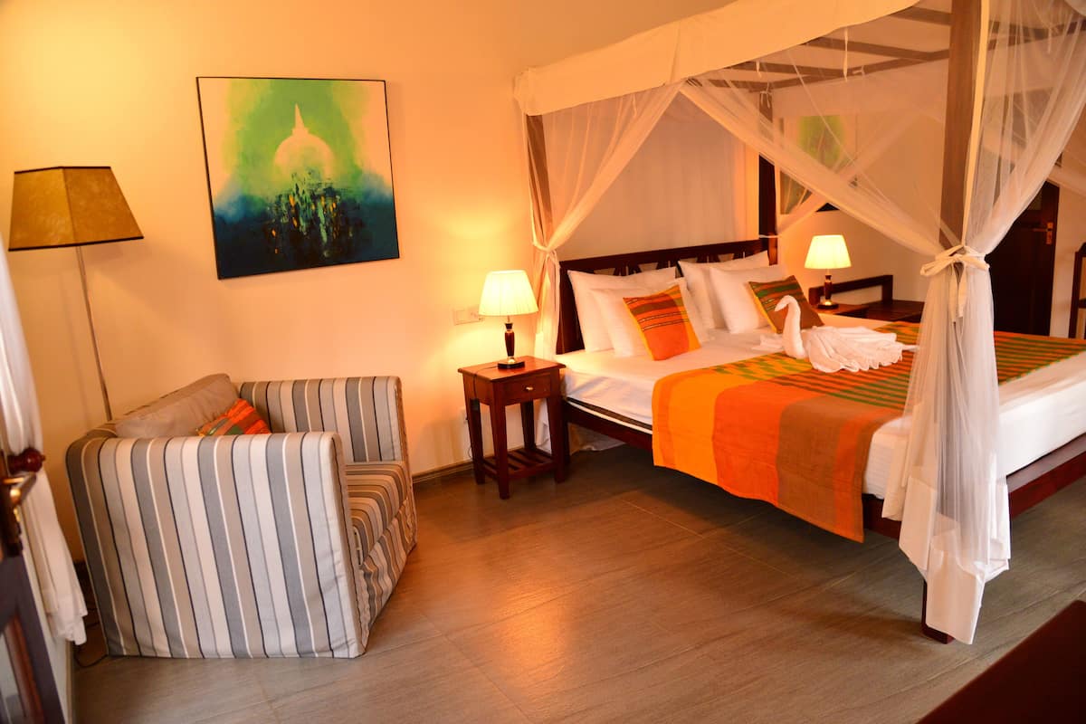 A hotel room with a four-poster bed, a striped armchair, a bedside table with a lamp, and a painting on the wall.