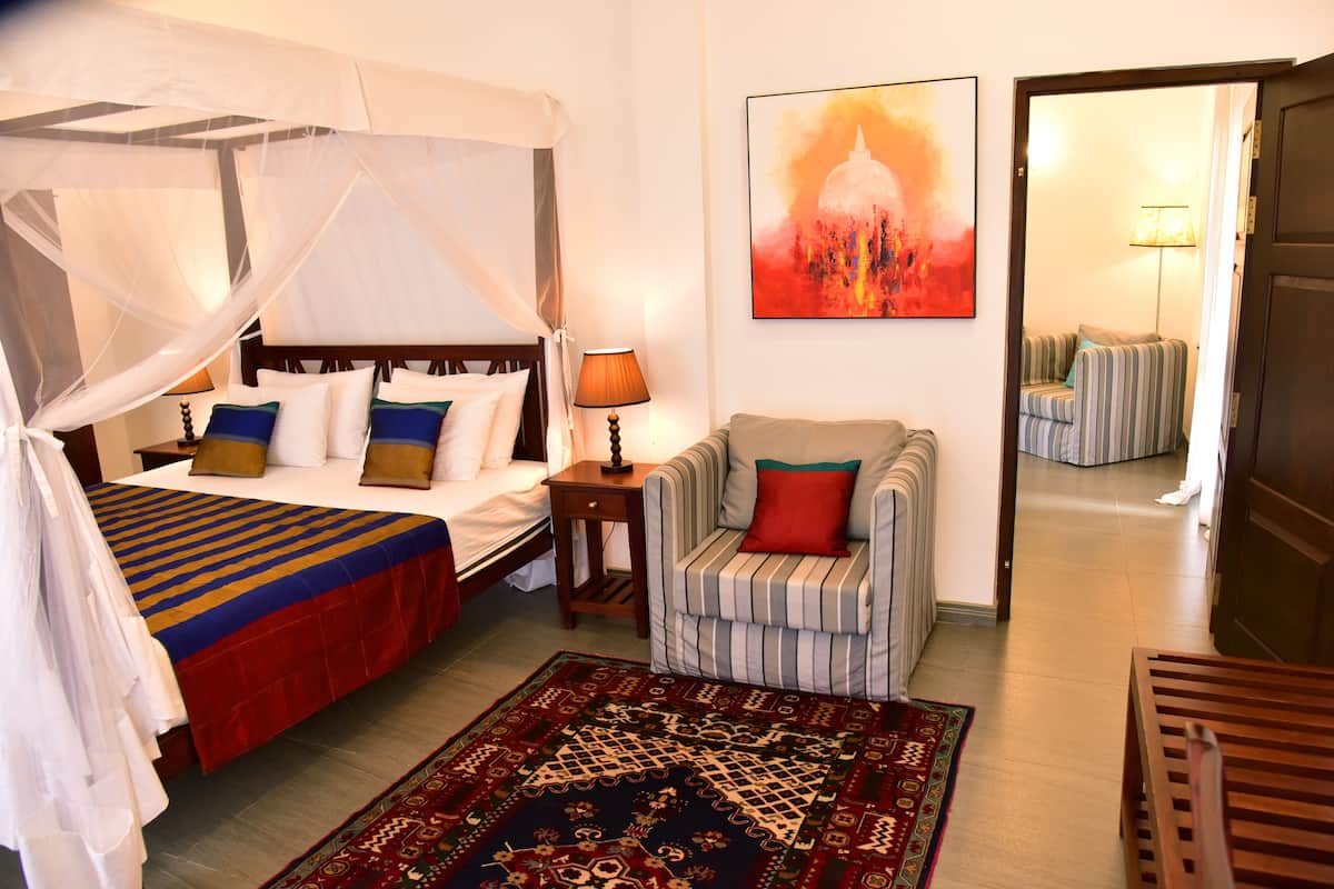 A hotel room with a four-poster bed, a striped armchair, a patterned rug, and a painting of a building.