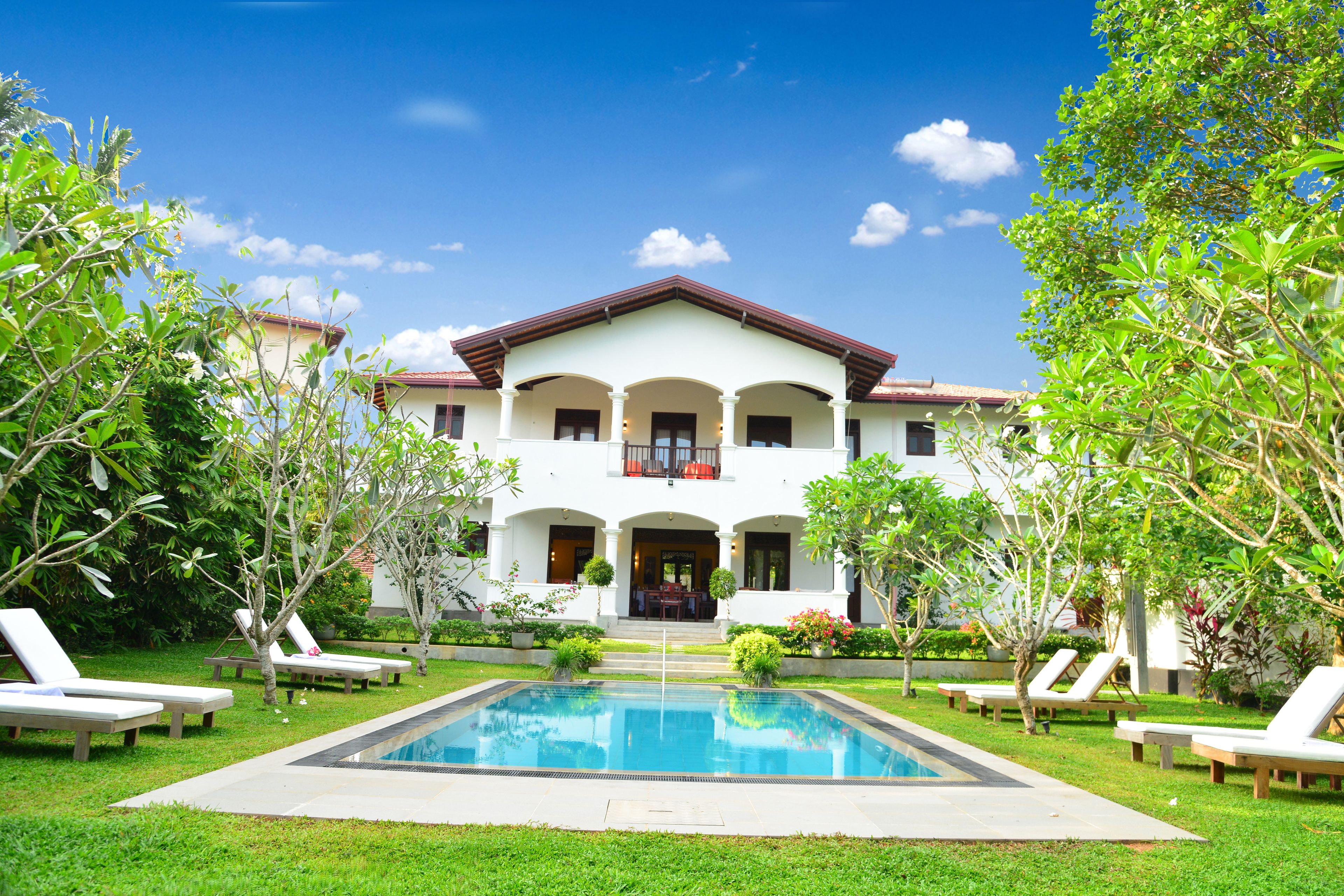 A two-story building with a swimming pool, surrounded by greenery and lounge chairs.