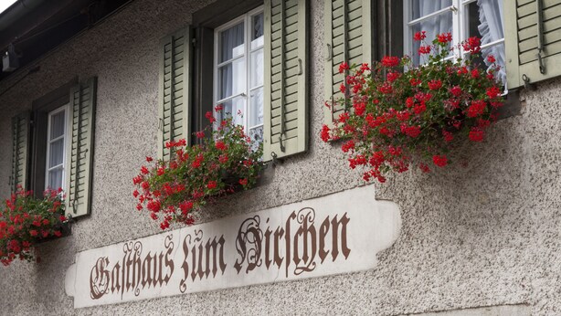 A building with a sign reading "Gasthof Zum Stuben" and flower boxes with red flowers on the windows.
