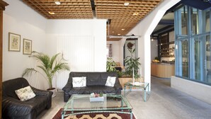 A living room with a black leather sofa, a glass coffee table, and a patterned rug. There are potted plants and a kitchen visible in the background.