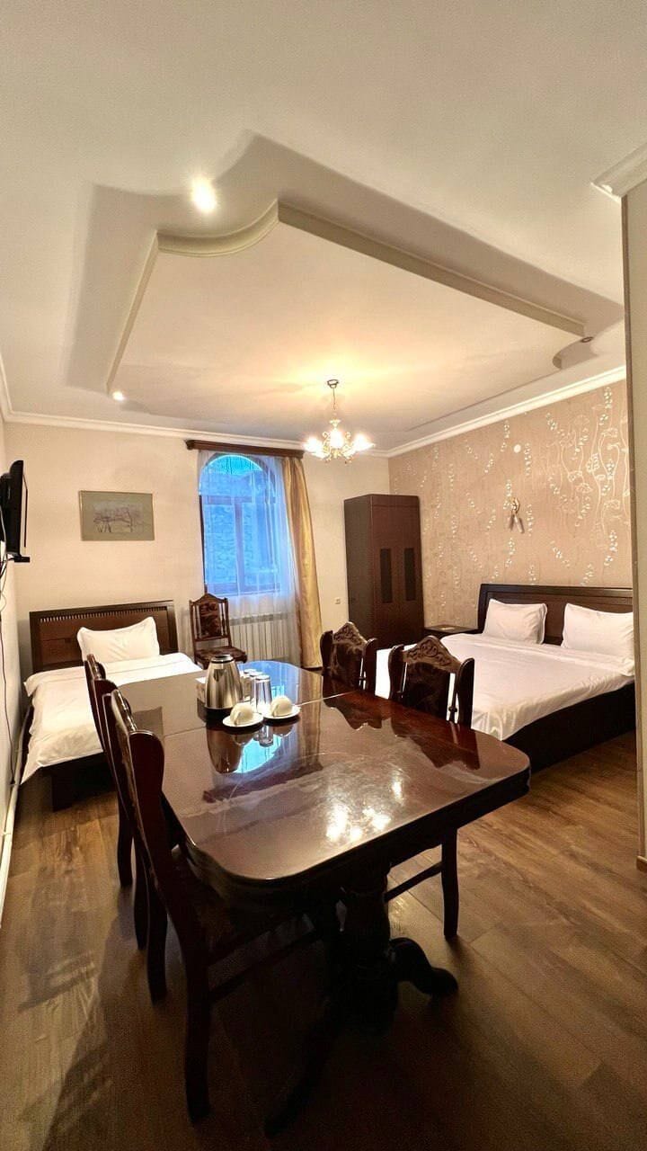 A hotel room with a dining table, two beds, a chandelier, and a window with curtains.