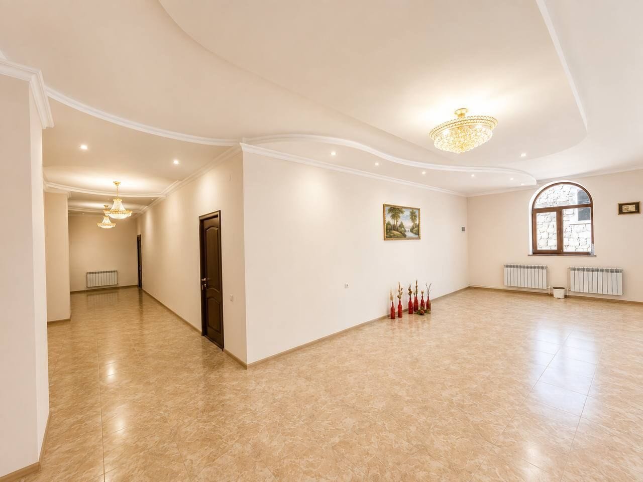 Um quarto amplo com teto alto, um candelabro, uma porta, um radiador e uma janela.