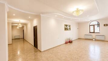 Um quarto amplo com teto alto, um candelabro, uma porta, um radiador e uma janela.
