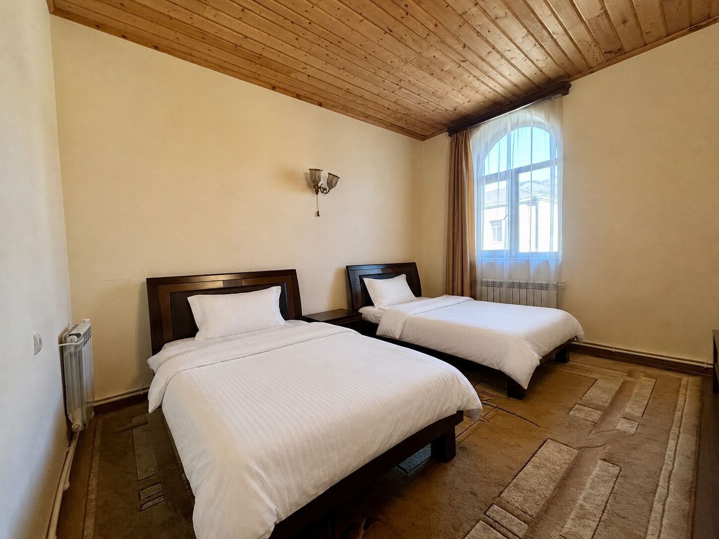 Habitació d'hotel amb dos llits individuals, sostre de fusta, radiador i finestra amb cortines.