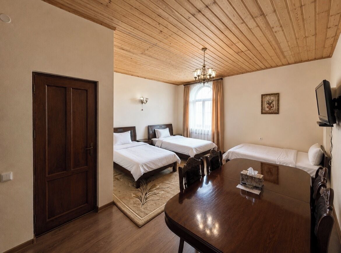 A hotel room with a wooden ceiling, two single beds, a sofa, a dining table, and a TV.