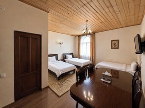A hotel room with a wooden ceiling, two single beds, a sofa, a dining table, and a TV.