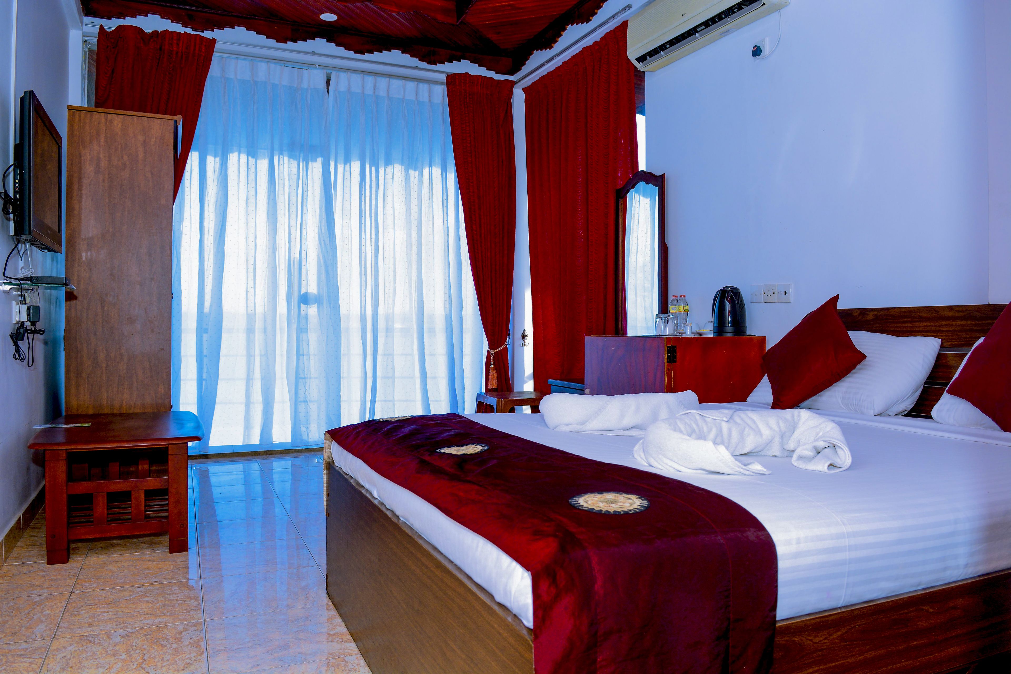 A hotel room with a large bed, red curtains, a wooden nightstand, a desk, and a mirror.