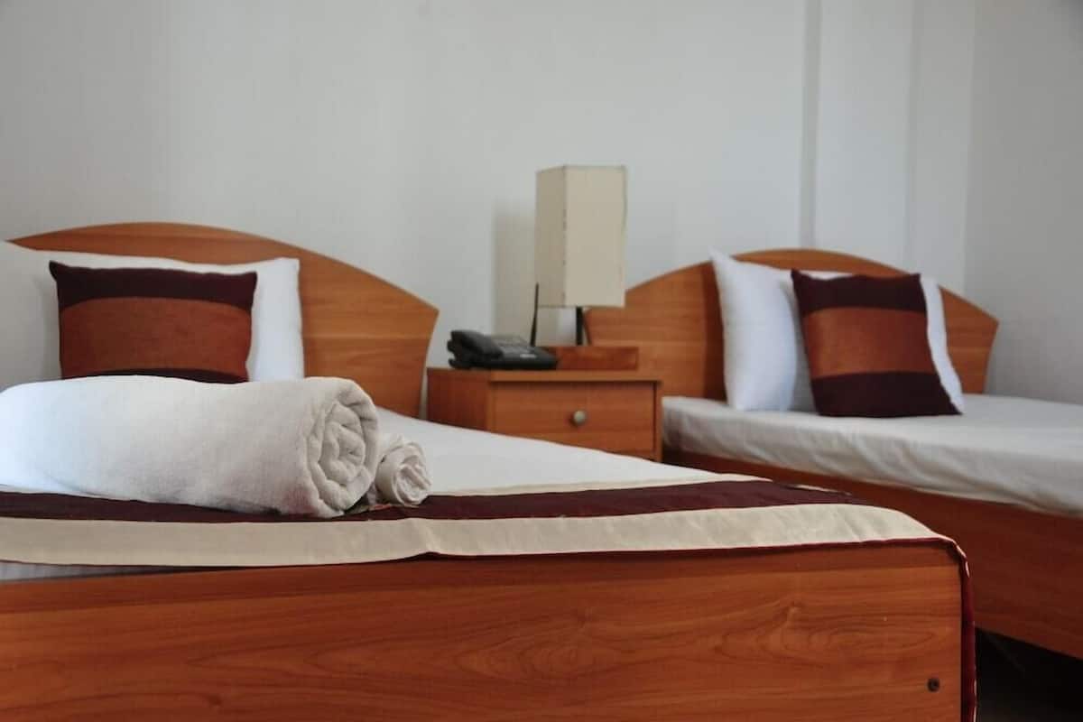 Ein Hotelzimmer mit zwei Betten, Holz-Kopfteilen, einem Nachttisch mit Lampe und einem auf dem Bett liegenden gefalteten Handtuch.