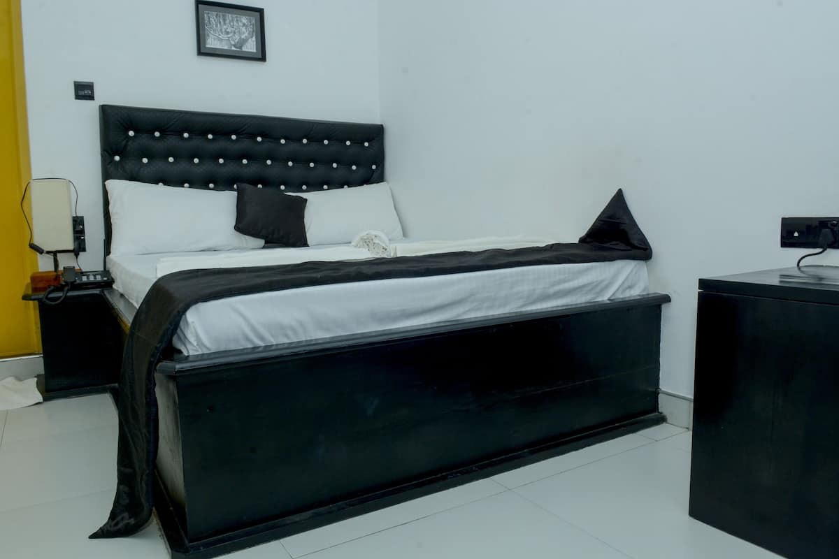 Ein ordentlich gemachtes Bett mit schwarzem Kopfteil, weißer Bettwäsche und einer schwarzen Decke. Ein Nachttisch mit Lampe und eine Steckdose an der Wand sind vorhanden.