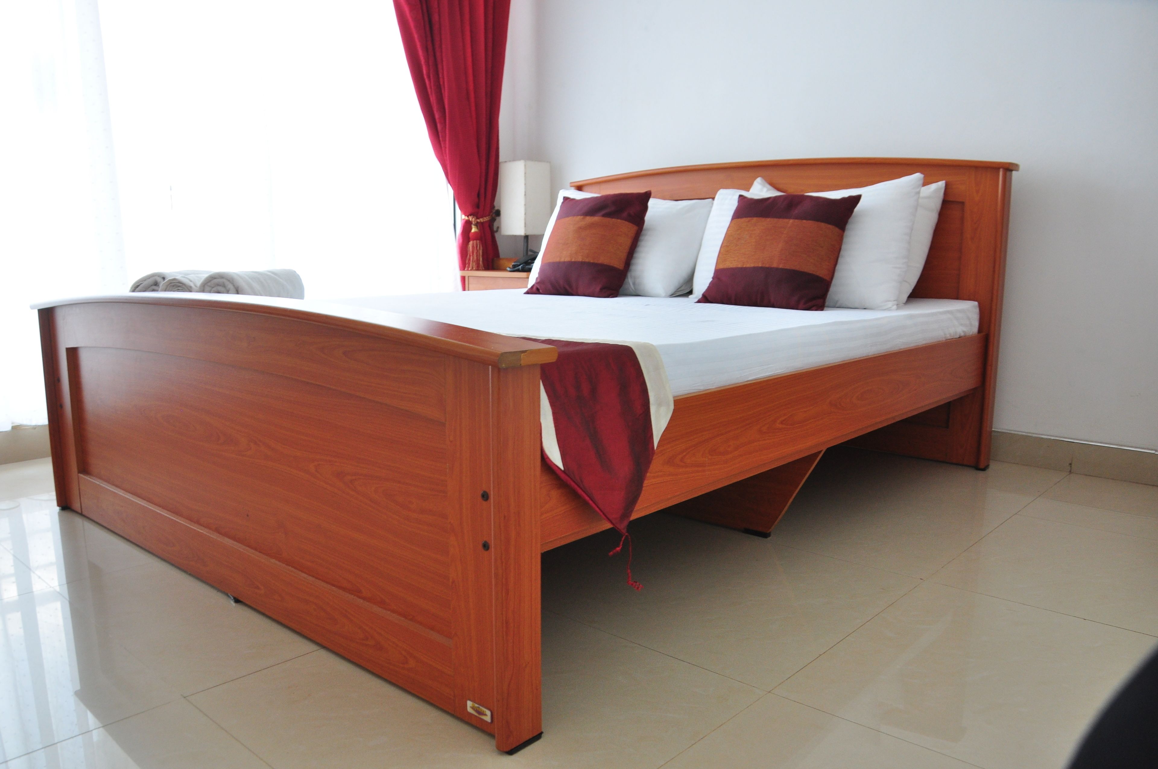 Una cama de madera con colchón blanco y almohadas rojas y marrones en una habitación con suelo de baldosas y cortina roja.