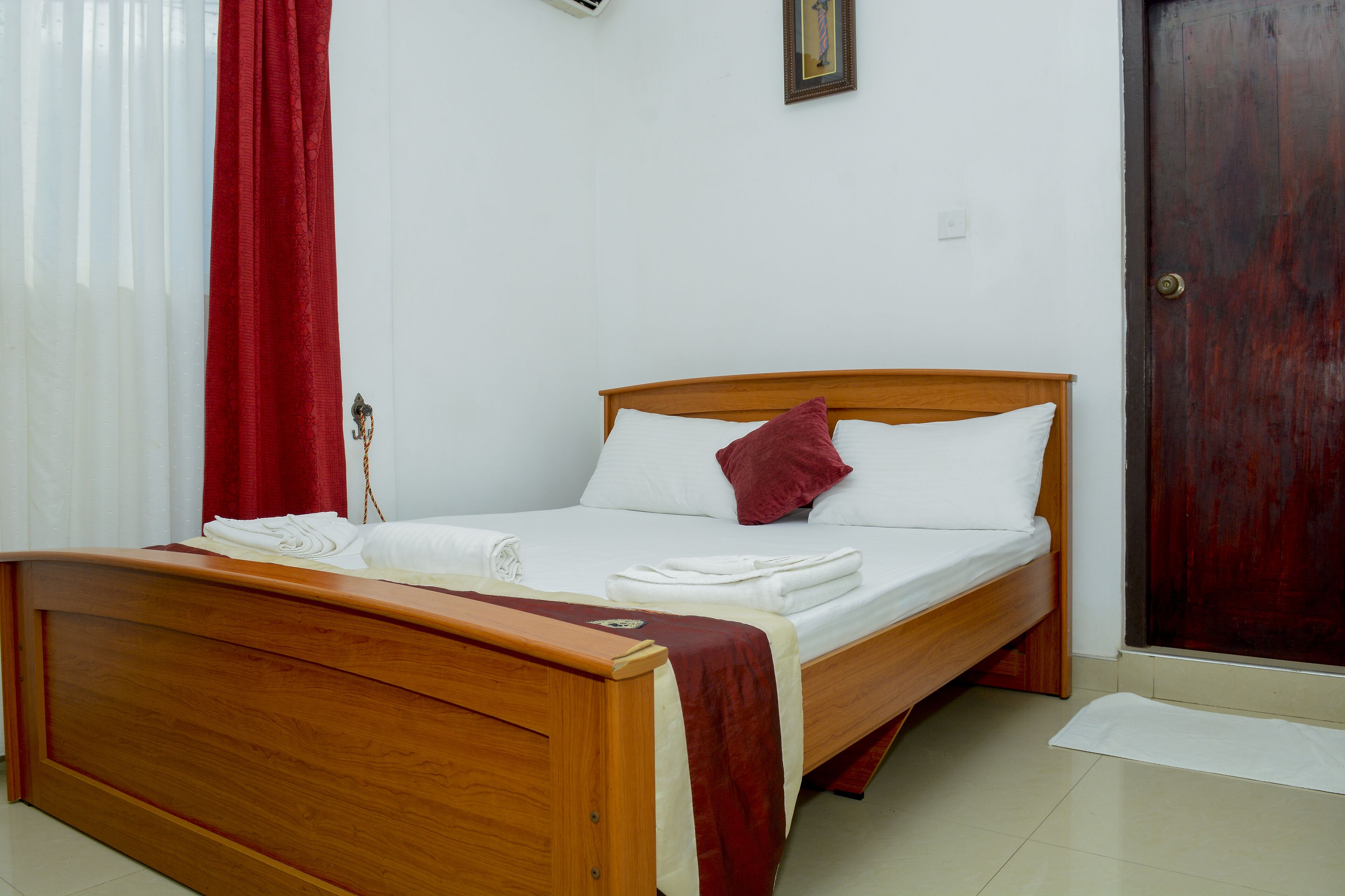 A neatly made bed with white and red bedding, a wooden headboard, and a red curtain on the window.