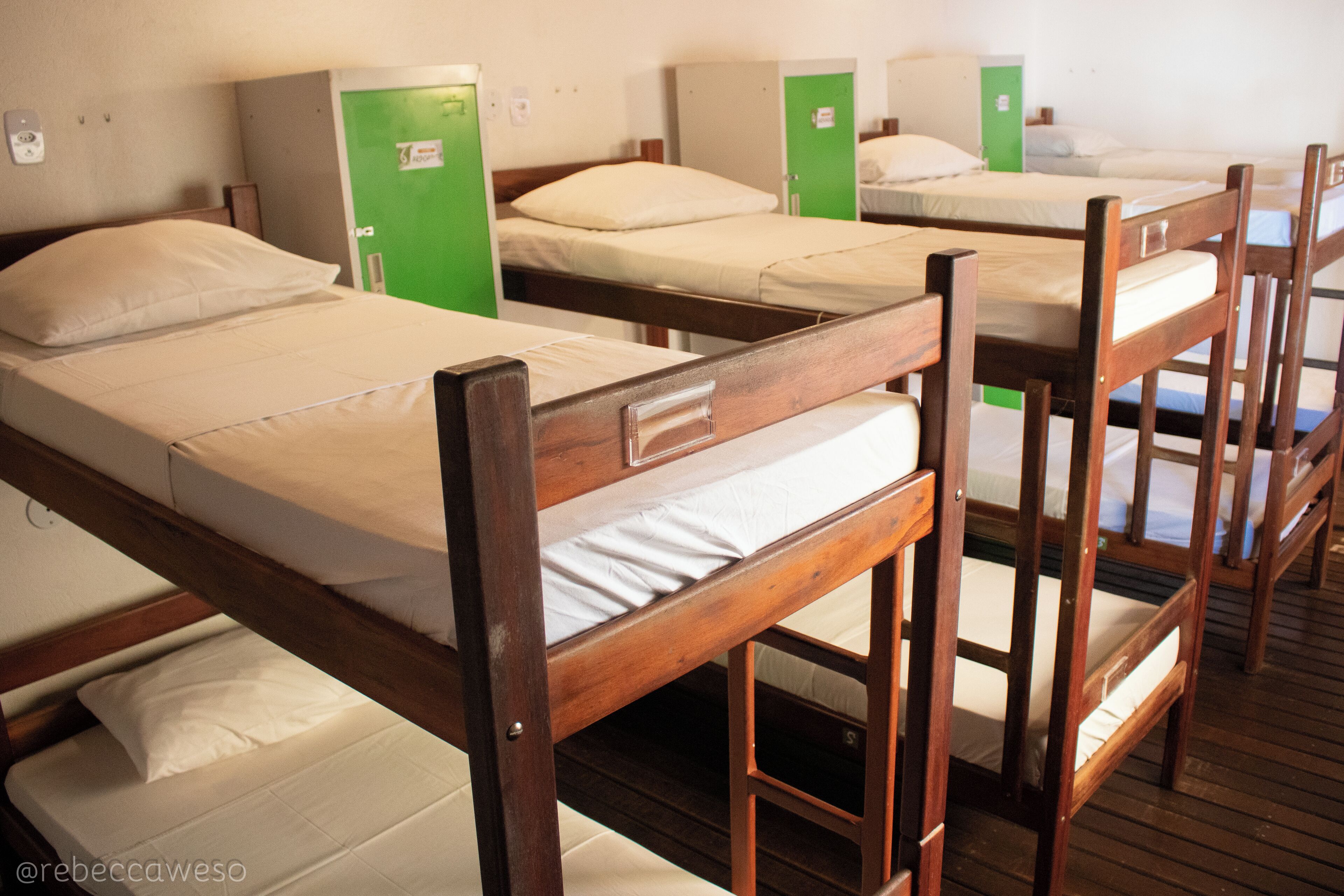 Una hilera de literas de madera con puertas verdes, cada una con colchón y almohada blancos.