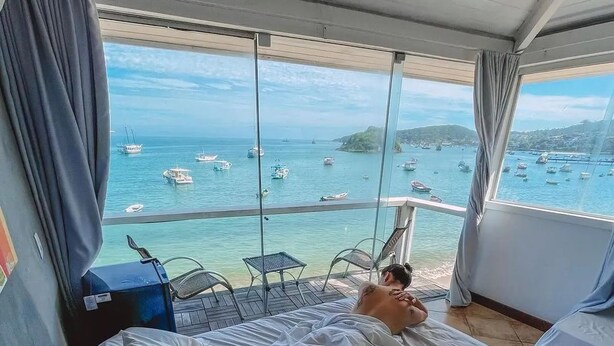 Een kamer met een bed, uitzicht op zee met boten en een balkon met stoelen.