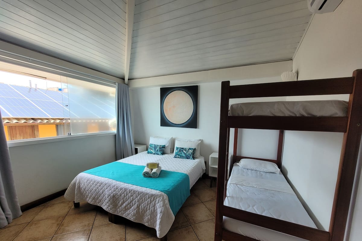 Una habitación con una cama, una litera, una ventana con cortinas, un reloj de pared y un techo con paneles solares.