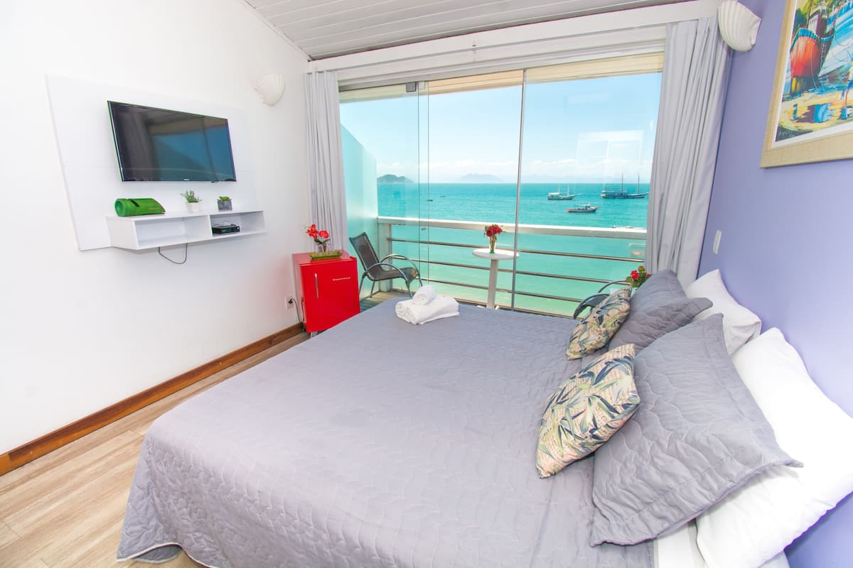 Habitación de hotel con una cama, un televisor montado en la pared, una mesita con un florero y vista al mar.