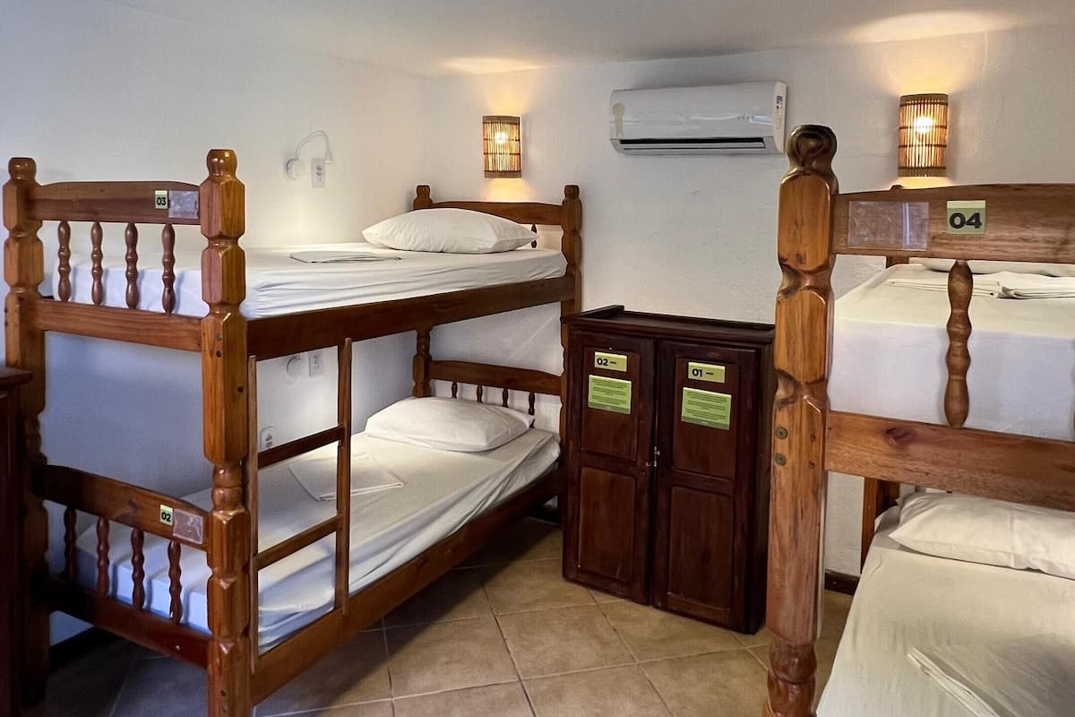 Una habitación con cuatro literas de madera, cada una con una etiqueta numérica. Hay una pequeña mesa con una lámpara, un equipo de aire acondicionado y una lámpara empotrada en la pared.