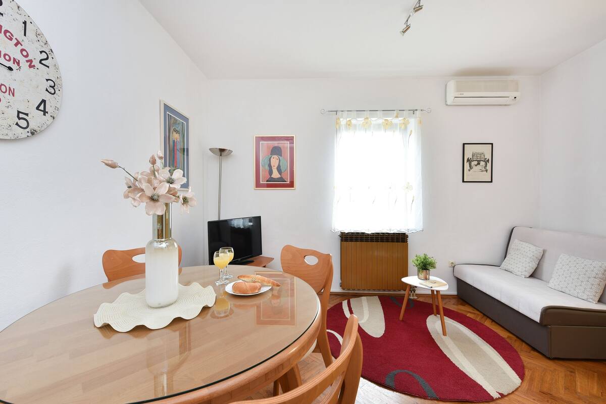 Una sala de estar con una mesa redonda de vidrio, un sofá, un televisor y un reloj en la pared.