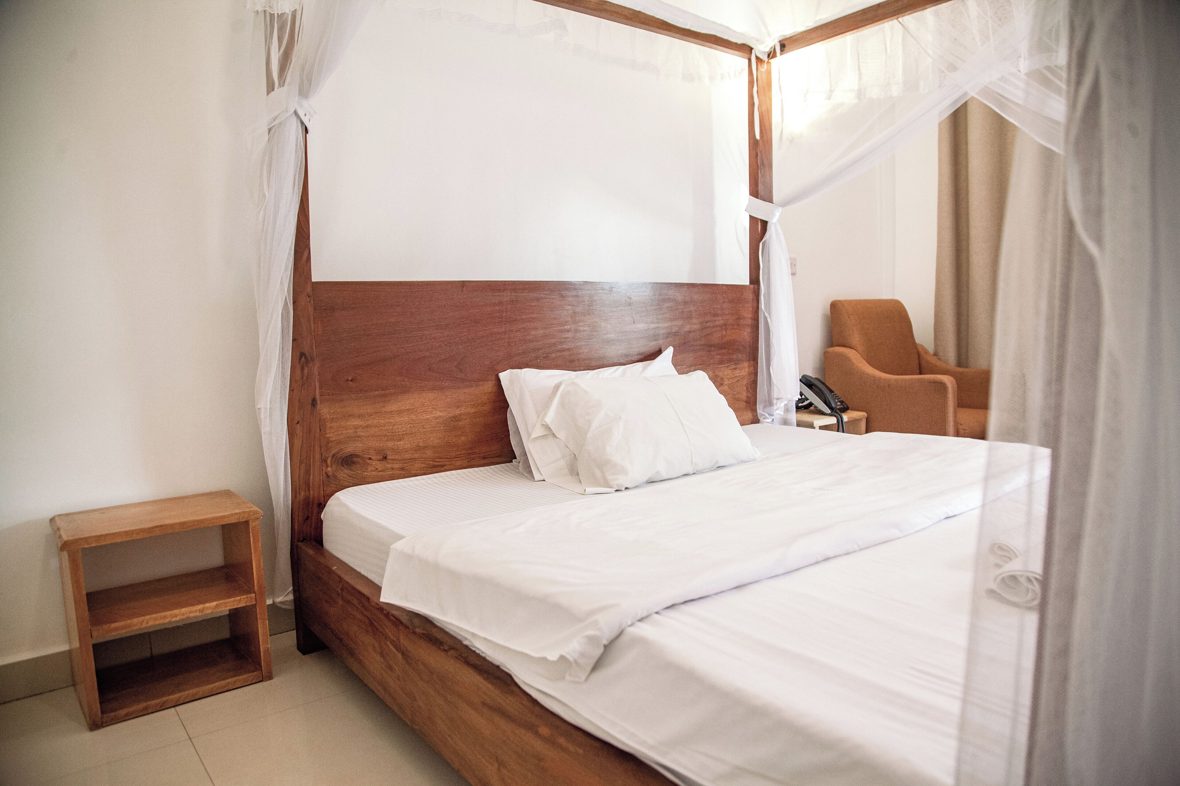 Una cama bien hecha con sábanas blancas, un cabecero de madera, una mesita de noche de madera y una silla en una habitación de diseño sencillo y limpio.