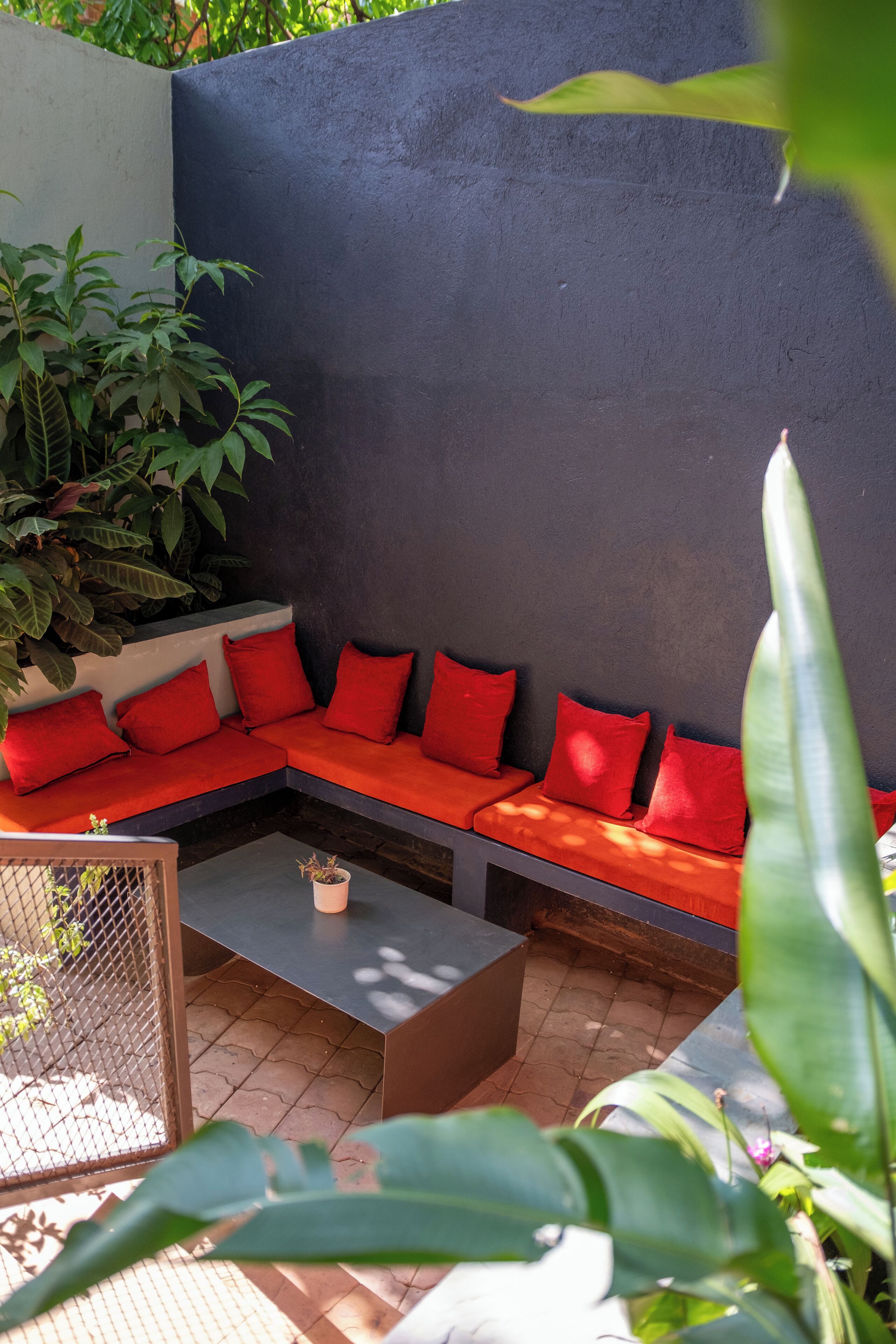 Un área de estar moderna al aire libre con pared oscura, cojines rojos y plantas en macetas.