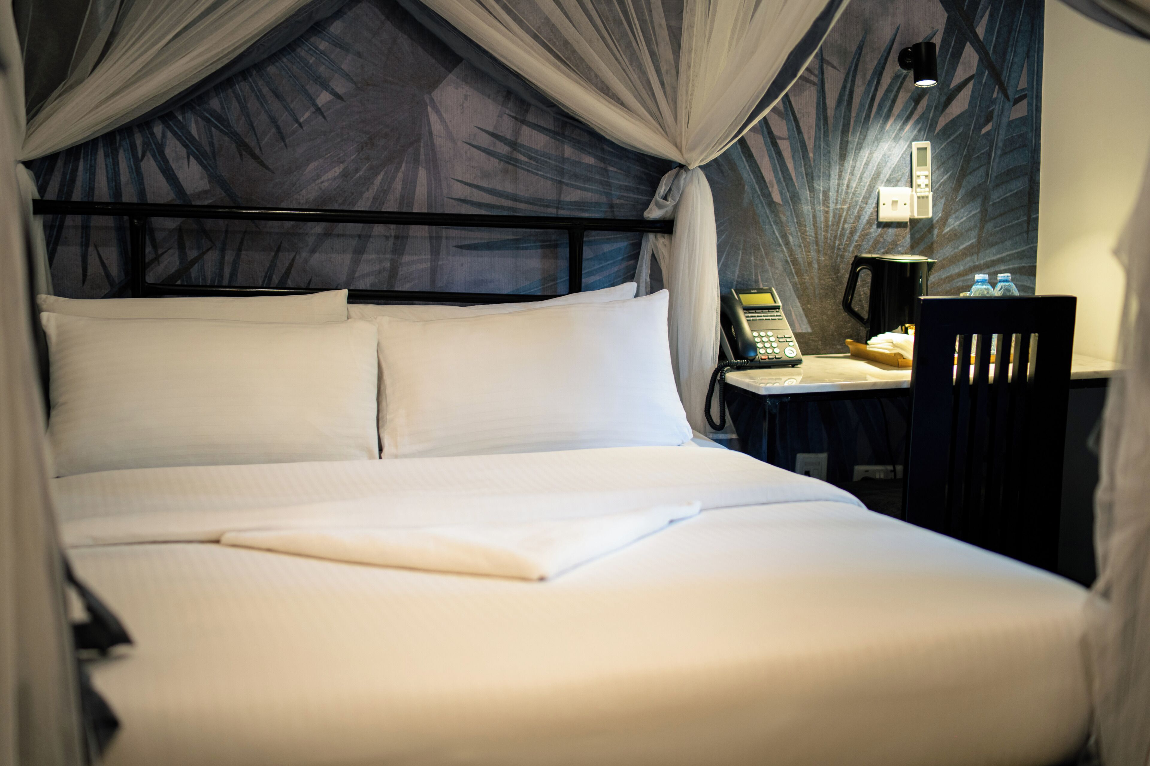 A neatly made bed with white linens and a canopy, a bedside table with a telephone and a kettle, and a chair.
