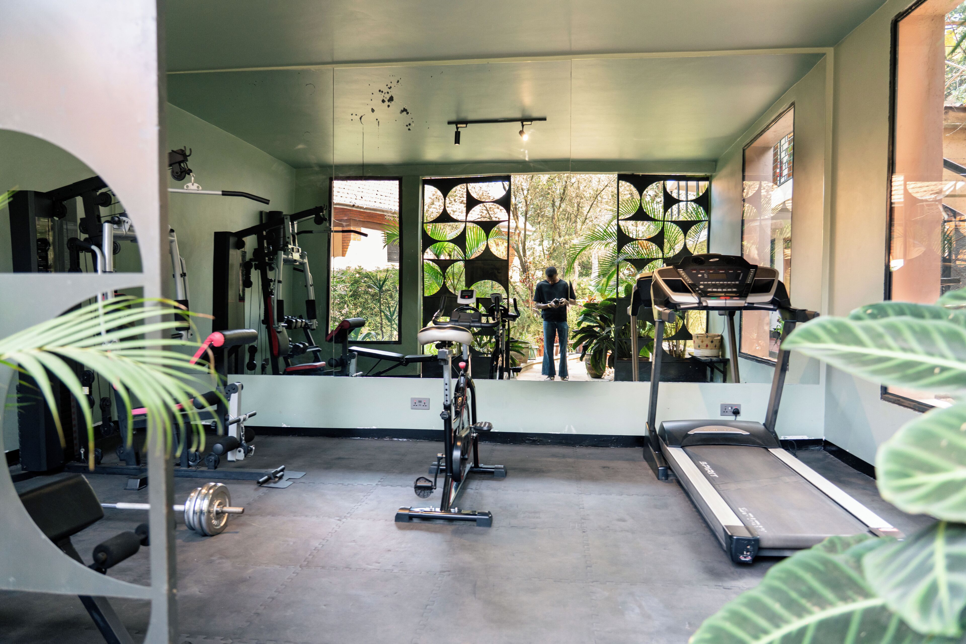 Ein Fitnessstudio mit Trainingsgeräten, Laufband und einem großen Fenster mit Blick auf Grünflächen.