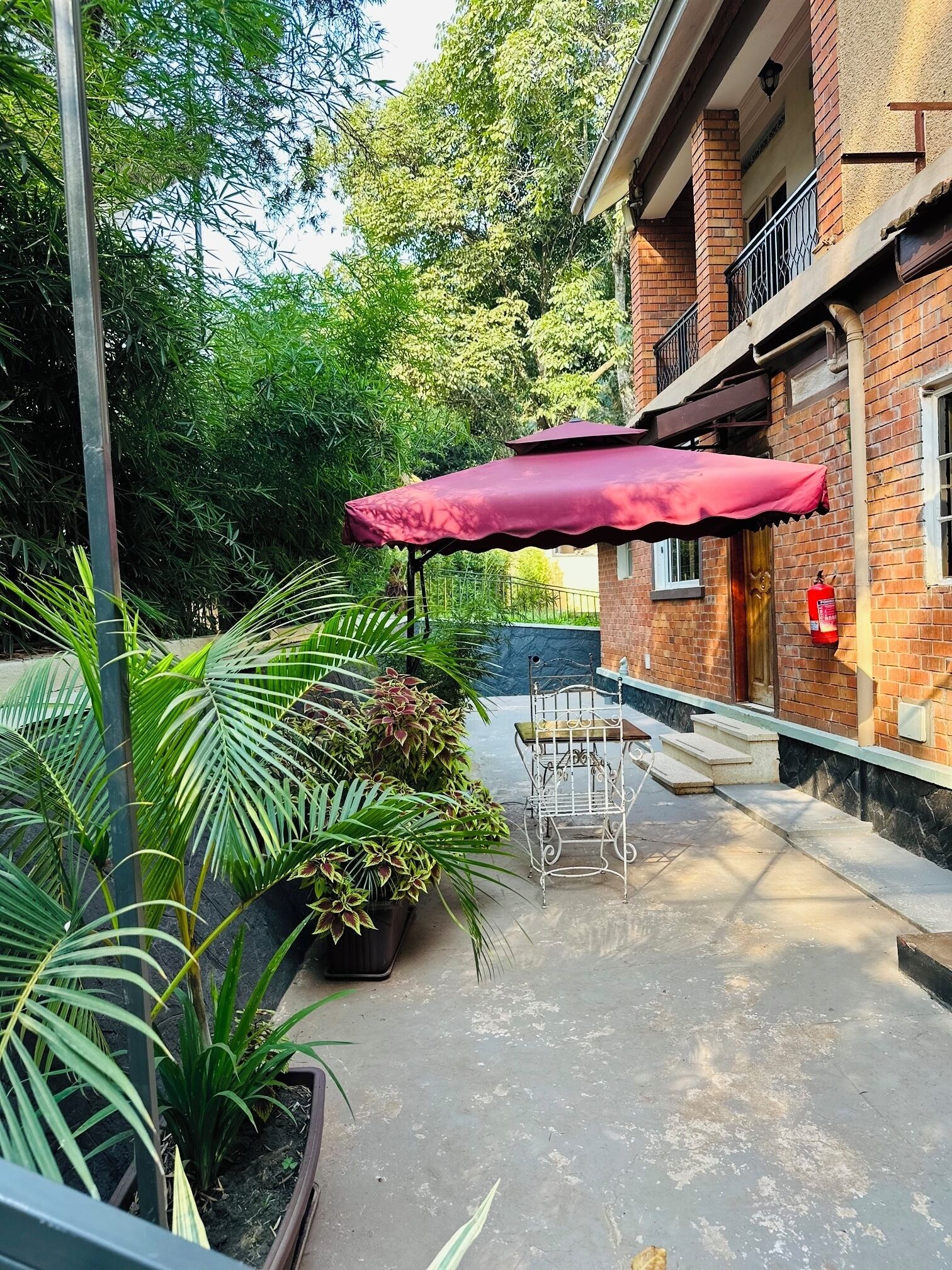 Área de pátio com um guarda-sol vermelho, plantas em vasos e um edifício de tijolos.