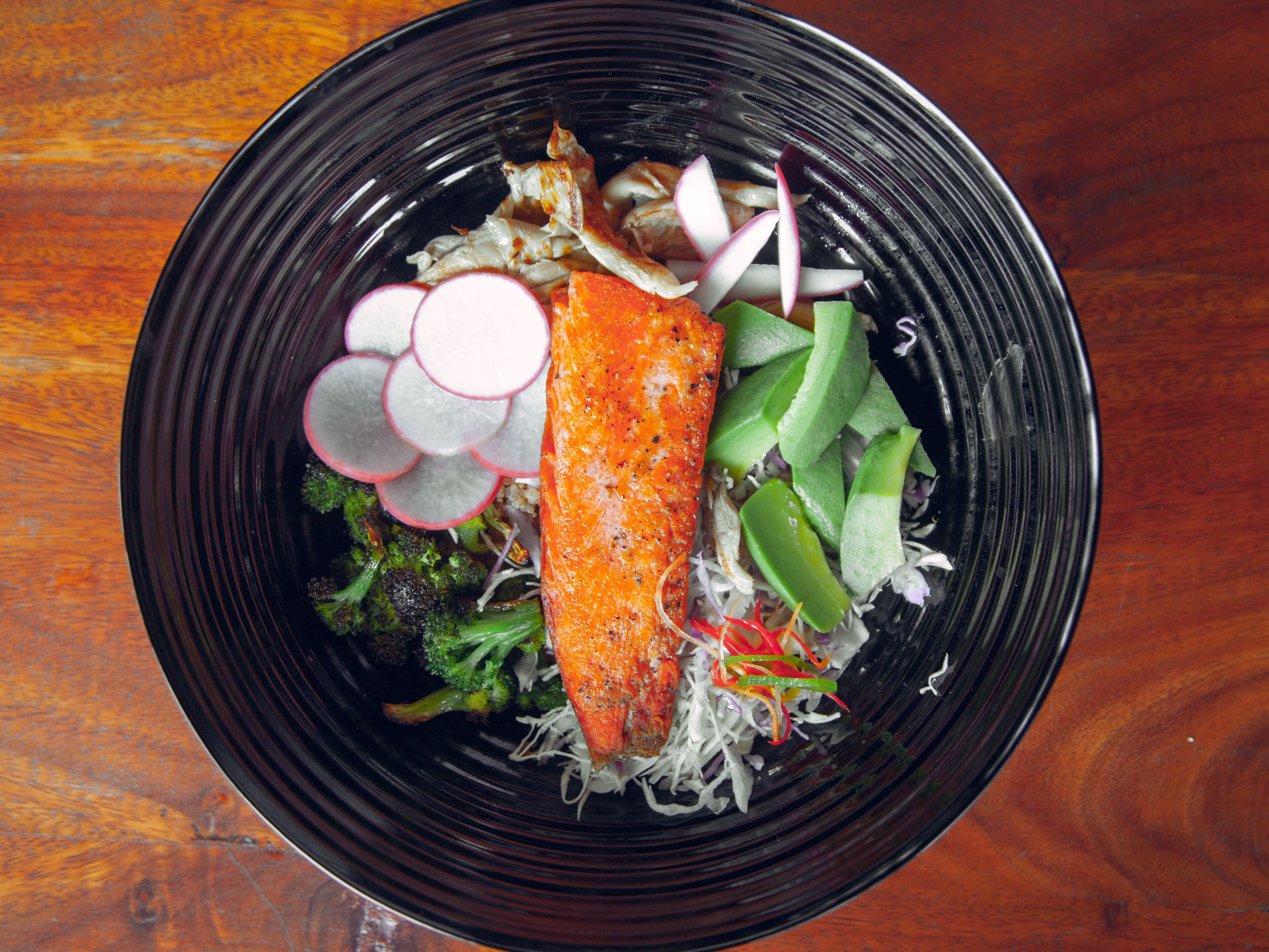 A black bowl containing salmon, radish slices, green vegetables, and shredded cabbage.