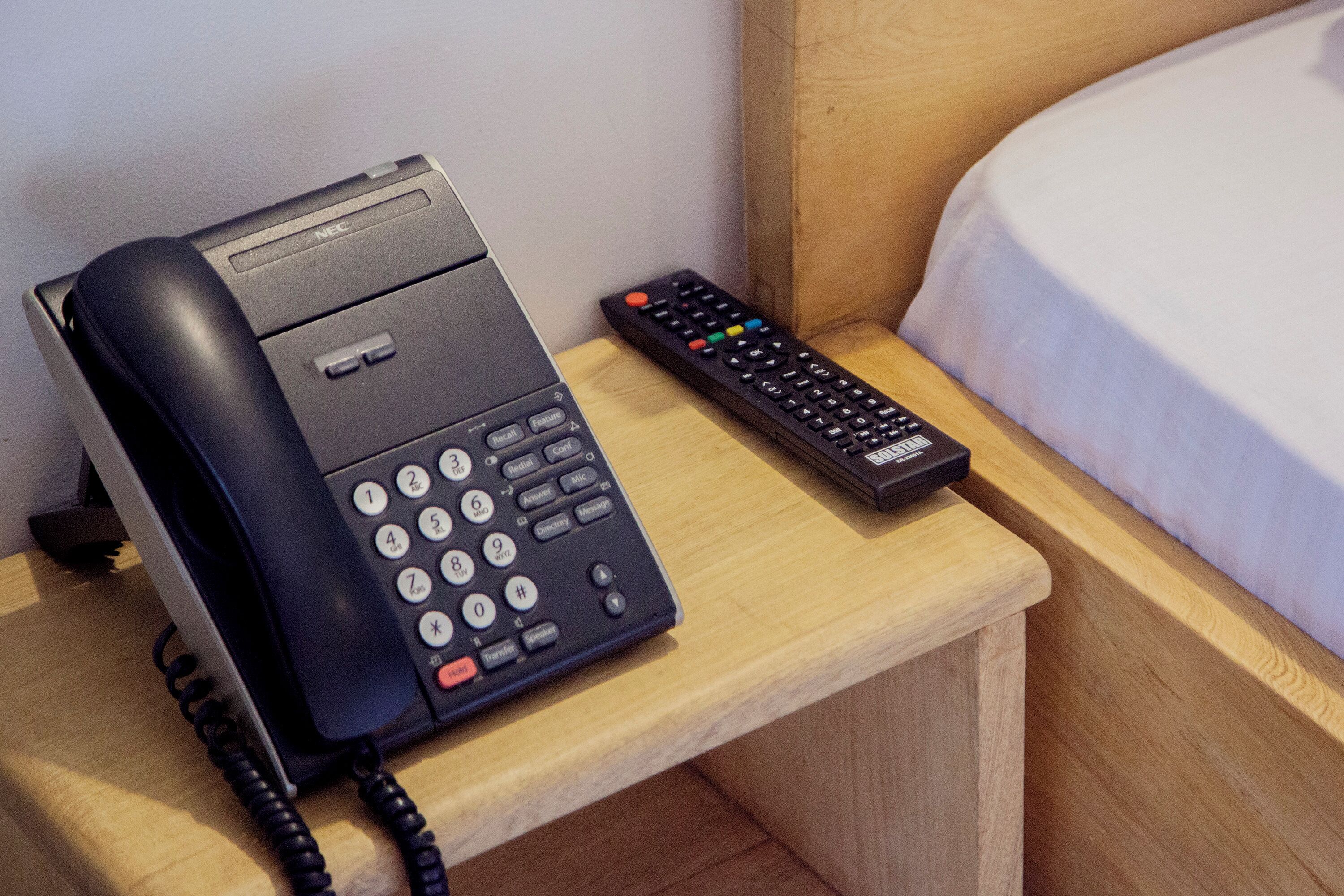 A hotel room with a bedside telephone and a remote control on a wooden nightstand.
