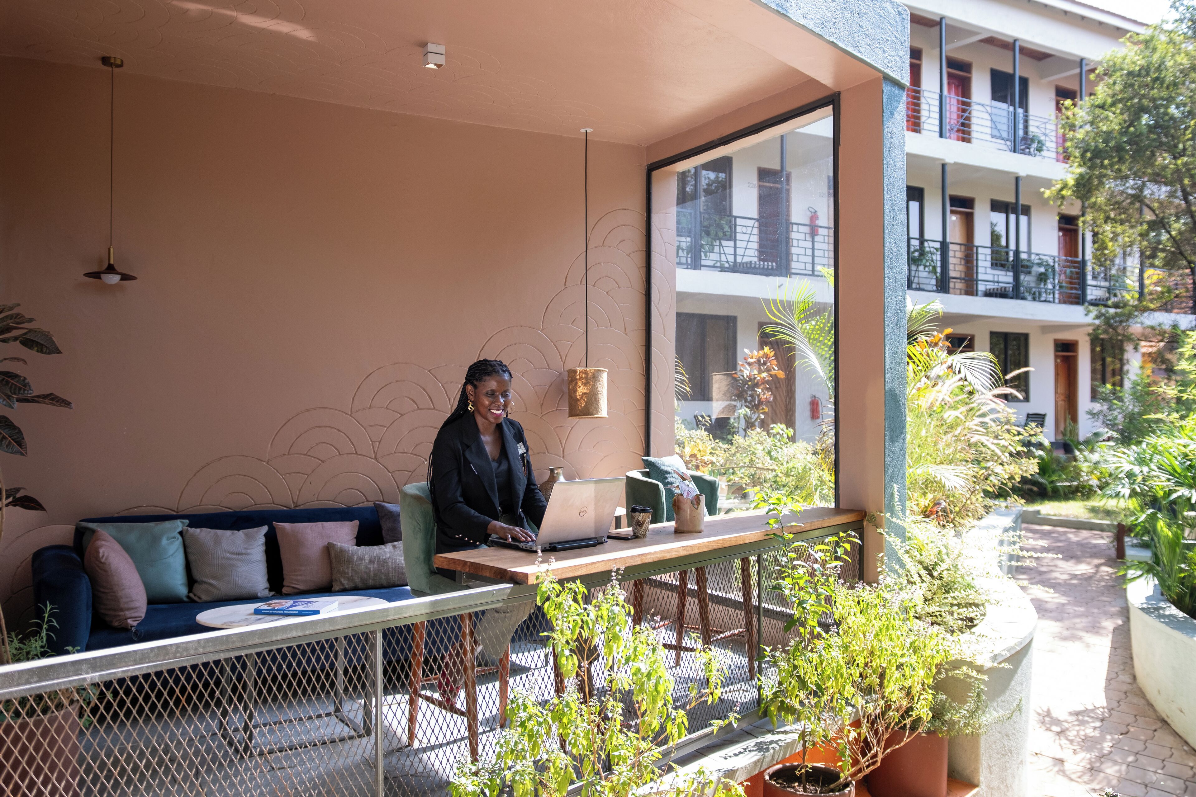 A person seated at a table with a laptop, a coffee cup, and a book on a bench in a sunlit outdoor area with a view of a building and greenery.