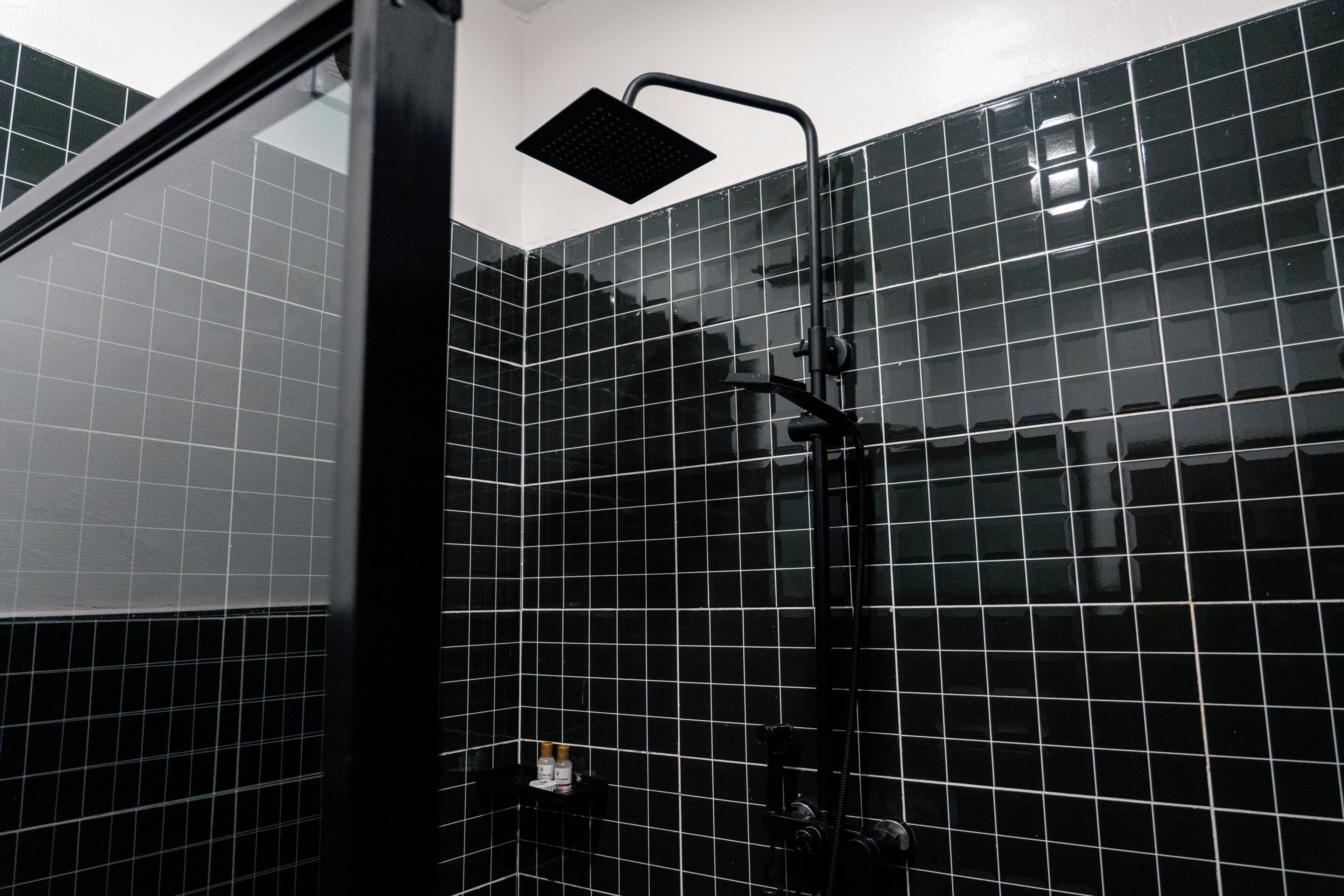 Un baño con paredes de azulejos negros, una ducha con pomo negro y una botella visible en el suelo.