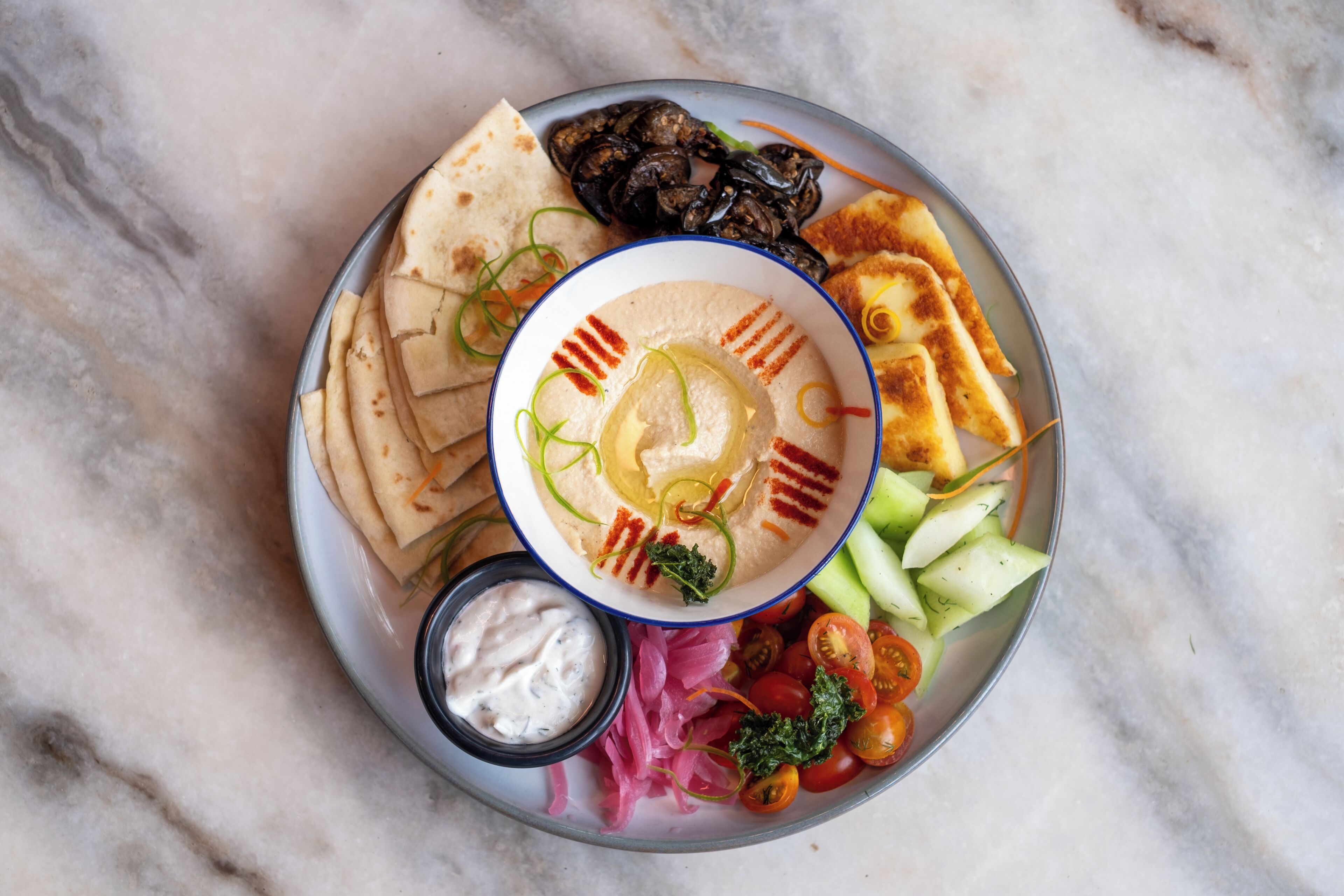A plate with hummus, pita bread, grilled vegetables, and a small bowl of sauce.