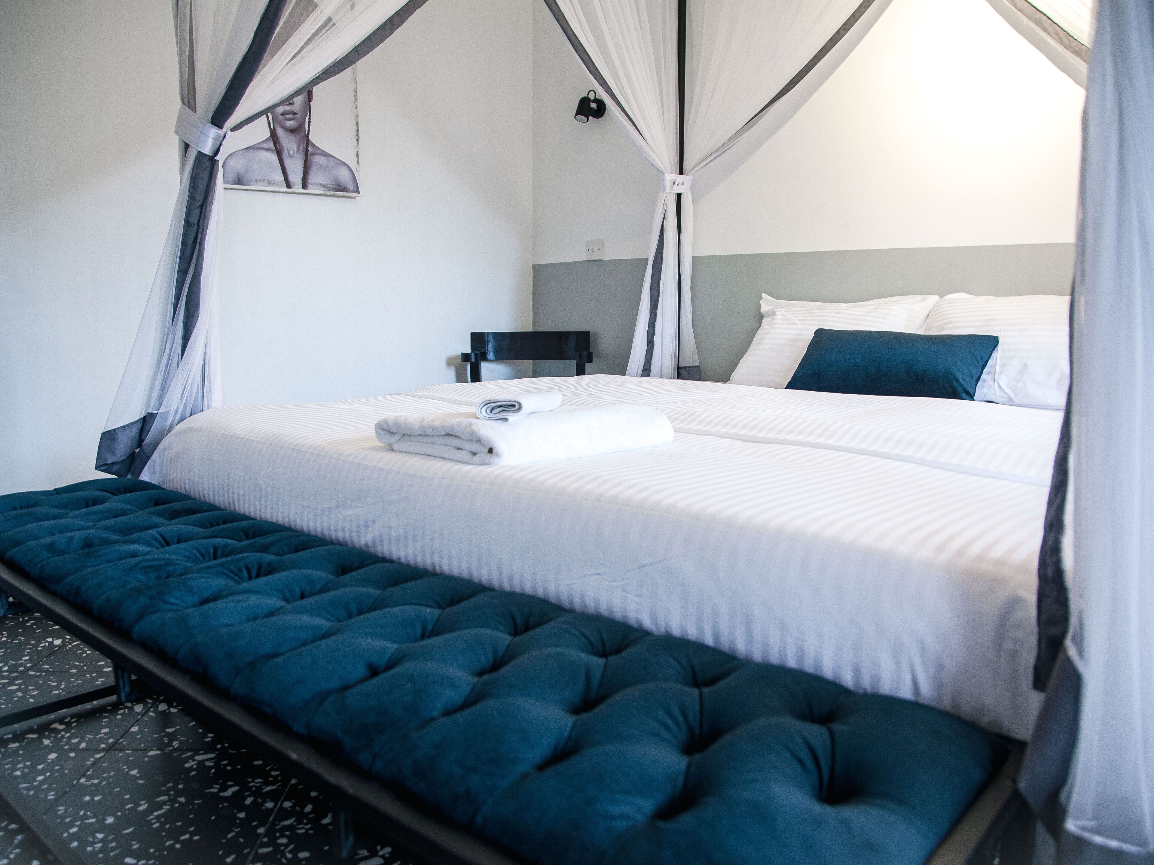 A neatly made bed with white linens and a blue accent pillow, a tufted blue bench, and a wall-mounted nightstand with a lamp.