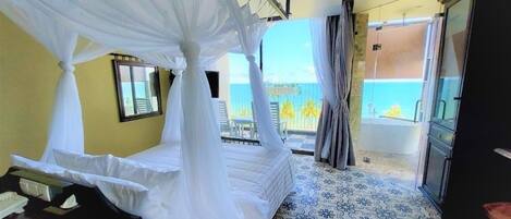 A room with a bed under a canopy, a bathroom with a bathtub, and a view of the sea.