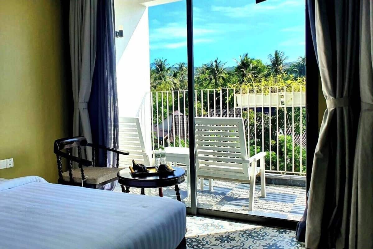 Une chambre d’hôtel avec un lit, une chaise, une petite table et un balcon offrant une vue sur les palmiers et les montagnes.
