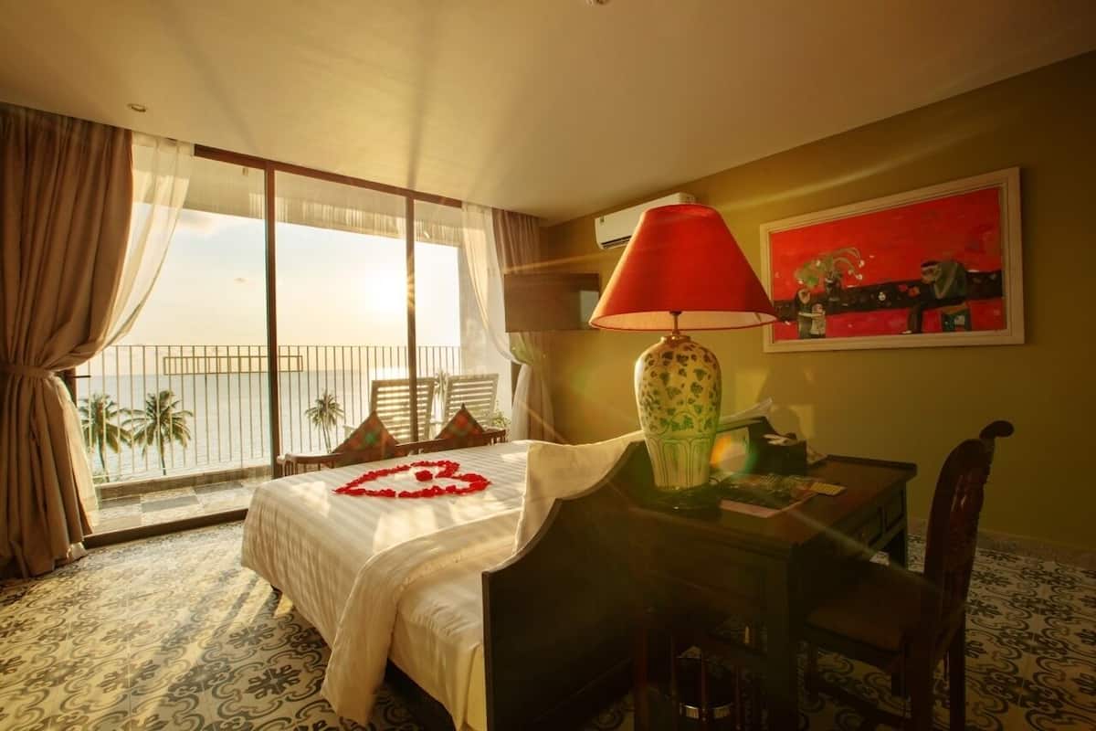 Une chambre d’hôtel avec un lit, une table avec une lampe, un tableau et un balcon donnant sur des palmiers.