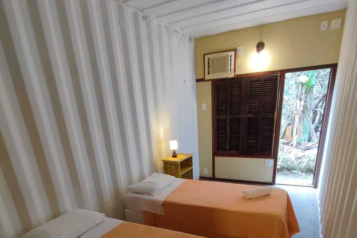 A room with two beds, each with a pillow and a blanket. There is a small table with a lamp, and a window with shutters.